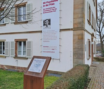 Zu sehen ist das Alleehaus mit einem Werbebanner für die Ausstellung und einer Informationsstele.